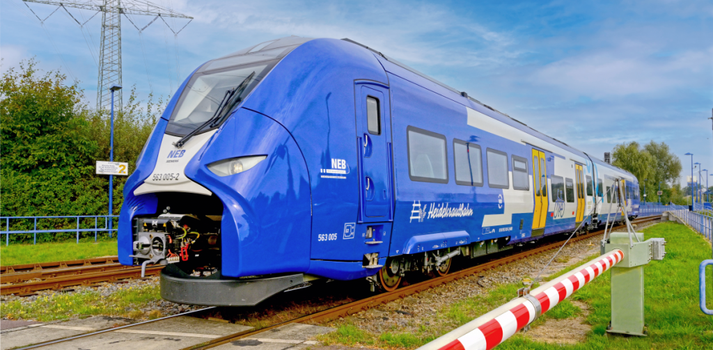 Blauer NEB Triebwagen der Heidekrautbahn an einem beschrankten Bahnübergang in lädnlicher Umgebung.