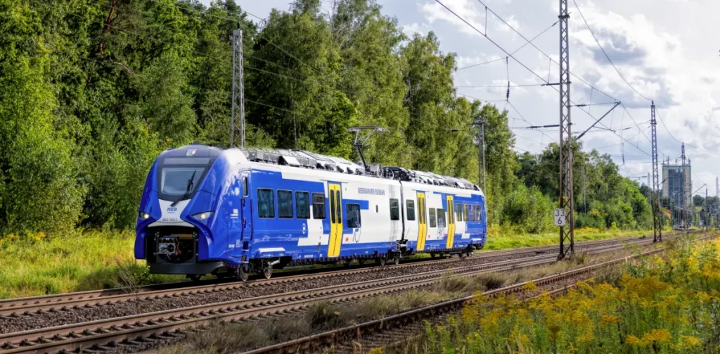 Blau-weißer Triebwagen der NEB Heidekrautbahn fährt auf einem Gleis mit Oberleitungen entlang an grünen Bäumen