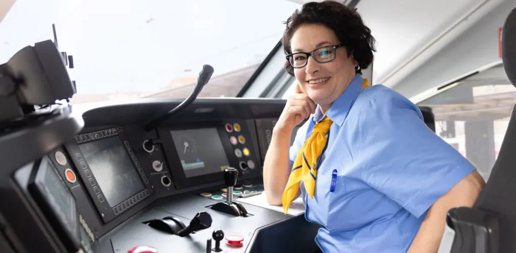 Freundlich lächelnde Triebfahrzeugführerin der NEB sitzt am modernen Führerstand eines Triebwagens und trägt dabei Dienstkleidung