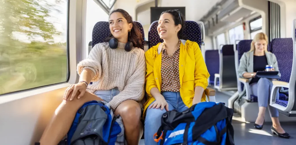 Zwei junge Frauen sitzen in einem sonnendurchfluteten Großraumabteil eines NEB Triebwagens, schauen nach draußen und lächeln. Im Hintergrund sitzt eine weitere Frau mit Kaffeebecher und Tablet-Computer.